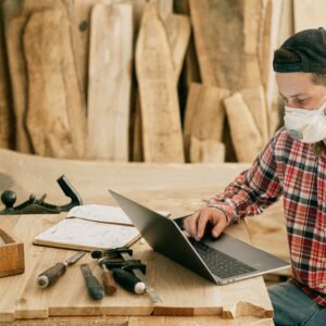 A craftsman wearing a mask, working on a laptop in a woodworking shop.